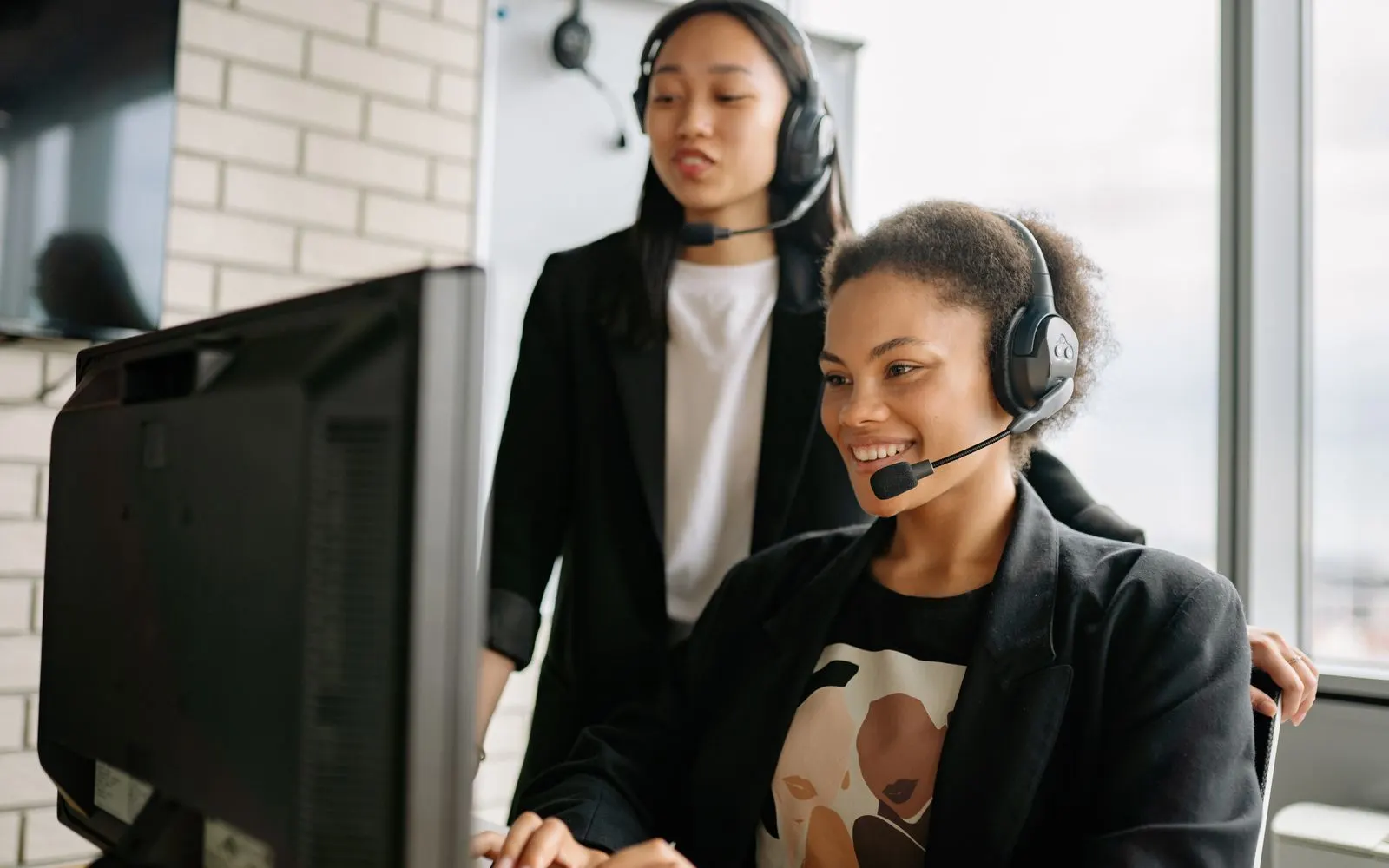 Two customer support agents with headsets smiling while helping customers at a computer