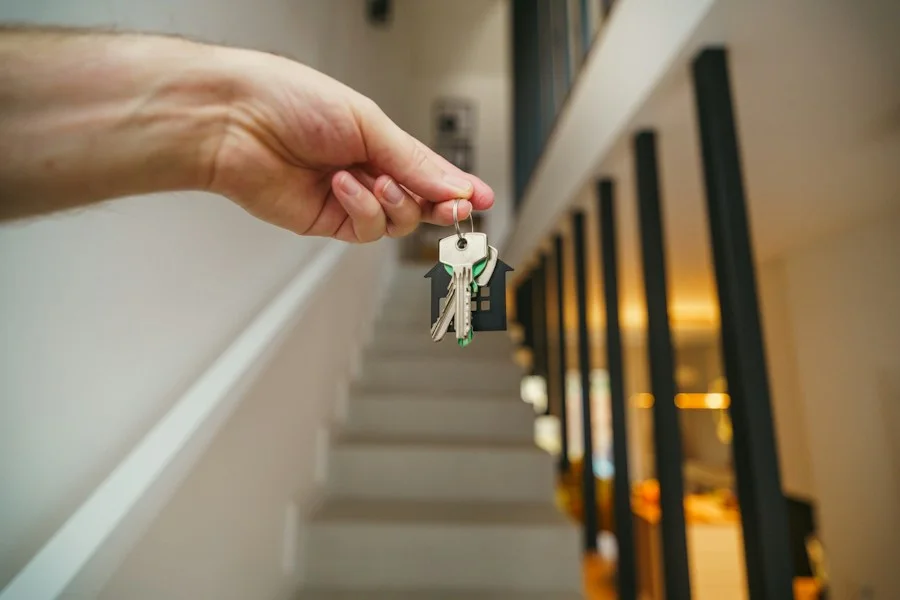 Hand holding keys with a house-shaped keychain in front of a staircase