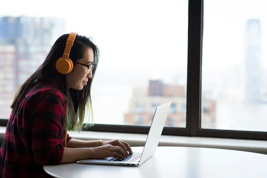 Woman with headphones working on a laptop beside a large office window