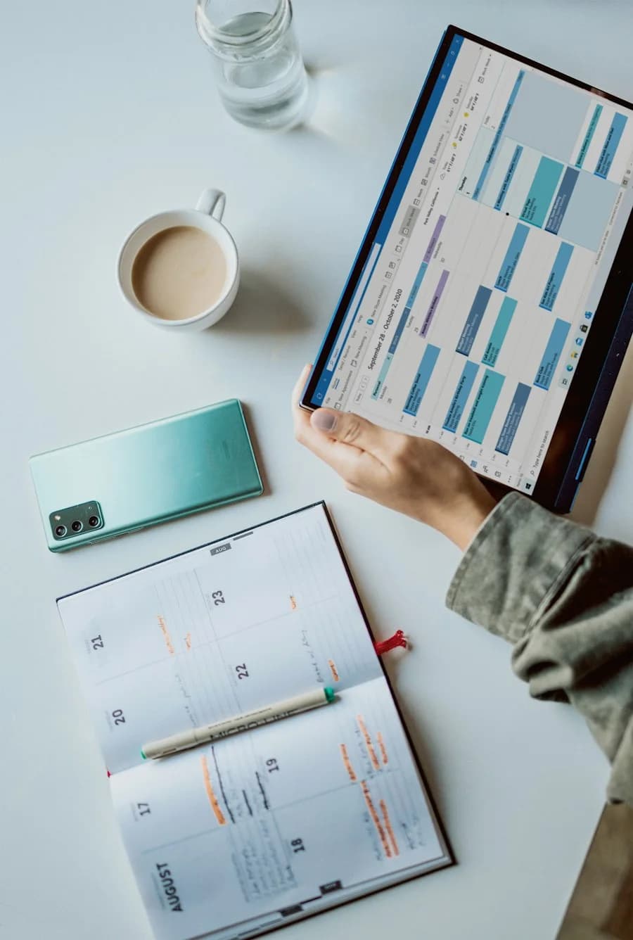 Hands holding a tablet with a calendar next to a paper planner and a cup of coffee
