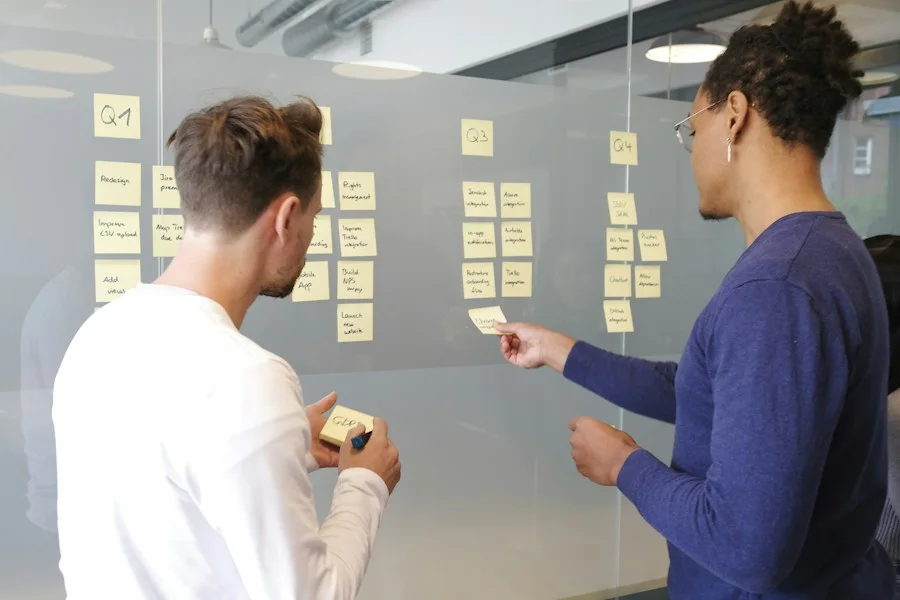 Two colleagues arranging sticky notes on a glass wall to plan a project