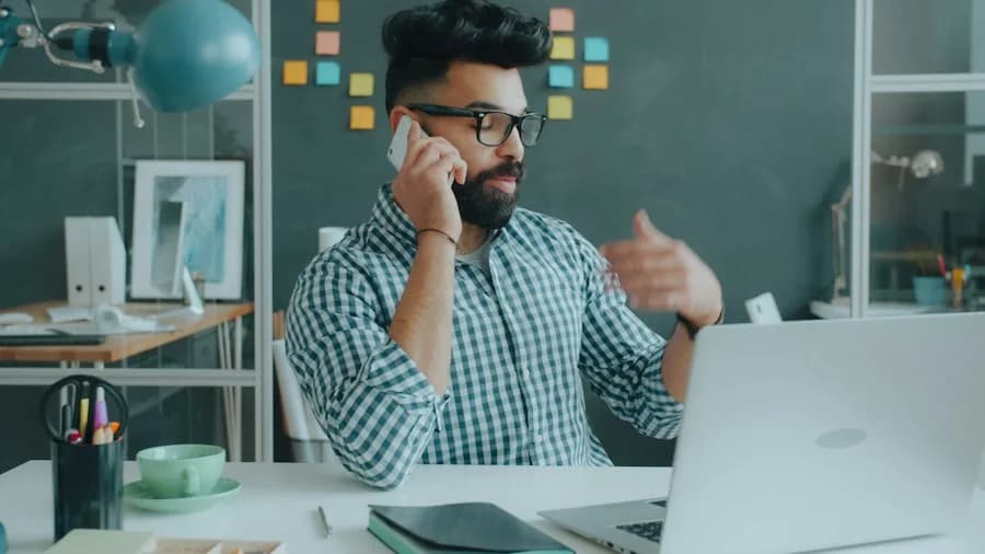 Man on a phone call at a laptop with sticky notes on the wall behind him