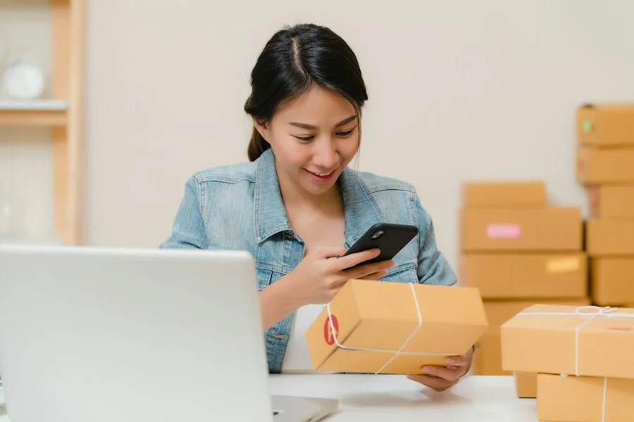 Woman scanning a parcel with her phone beside a laptop and boxes